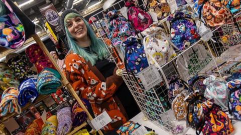 Becky Forster is standing behind her That's Sew Becky stall which offers clothing and accessories. T-shirts and bags are laid out, all in bright colours. Ms Forster has dyed light green hair and is wearing a black woolly hat and smiling. The picture has been taken at an angle.