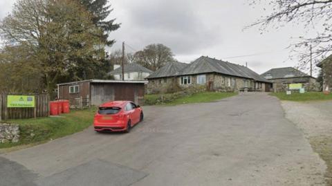 Google Street View image of YHA Dartmoor youth hostel. A red Ford Focus is driving into the driveway area of the site. Several stone buildings and a wooden shack are at the site.