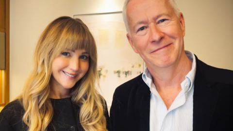 Jennifer Lawrence and John Wilson stand side by side and smile into the camera.