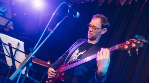Simon Gall in front of a microphone playing guitar. He is wearing glasses and a blzer style jacket over a t-shirt. 