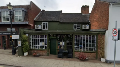 The bar is nestled between other buildings and is painted olive green with a low roof and grid windows and some seating outside with street greenery.