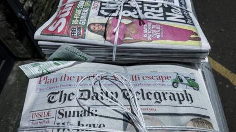 Bundles of newspapers tied with string, featuring The Sun with a headline about Charles and Harry's reunion, and The Daily Telegraph with the headline 'Sunak: We have everything to fight for.'