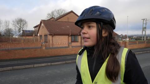 A young girl with long dark hair stood at the side of a road with a black cycle helmet and green hi vis jacket