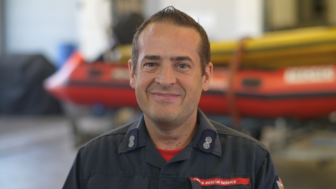Matthew Oxborrow a member of Norfolk Fire and Rescue Service. He is smiling and looking directly at the camera. He is wearing his fire service uniform and behind him is a boat used for rescues.
