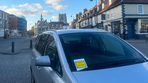 A square yellow parking ticket on the windscreen of a silver-coloured car parked on the paving stones of a market square with rows of shops and a church in the background