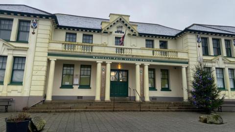 The image shows a Victorian-era building that is cream in colour and has a Christmas tree outside it.