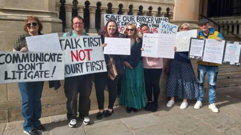 A group of around eight people stand outside a stone building with columns, holding handmade protest signs. The signs include messages such as “Communicate don’t litigate!”, “Talk first not court first,” and “Save our burger vans".