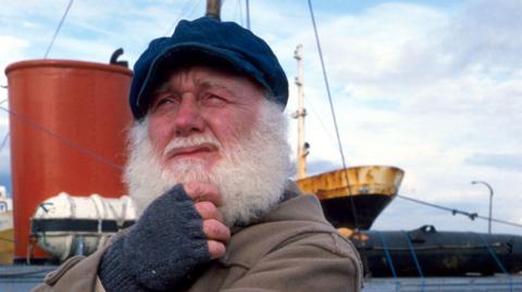 A bearded man with a blue cloth cap, grey fingerless gloves and a brown coat stands in front of a ship. He is holding his hand to his chin and looking pensive 
