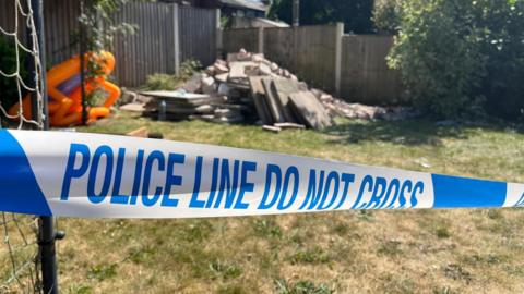 Police tape stretched across a garden lawn. A pile of rubble and debris can be seen in the garden.