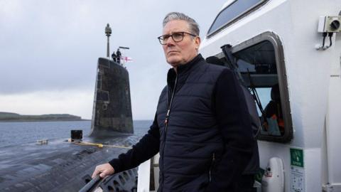 Keir Starmer on a boat next to a grey nuclear submarine. He is wearing a blue gilet over a blue or black jumper. He has grey hair and is wearing glasses. There are two men standing on the conning tower of the submarine