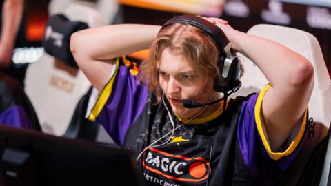A competitor at the Esports World Cup watches a screen intently with their hands clasped behind their head, creating an impression of anticipation or nervousness. They wear a large gaming headset with a microphone and a black and purple team jersey.