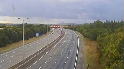 A traffic camera view of a motorway, with no cars visible on one carriageway. There are grass verges and trees on either side