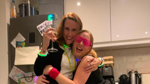 Two women embracing and laughing. They are hugging and wearing colourful accessories. One has a big glass of white wine in her hand. 