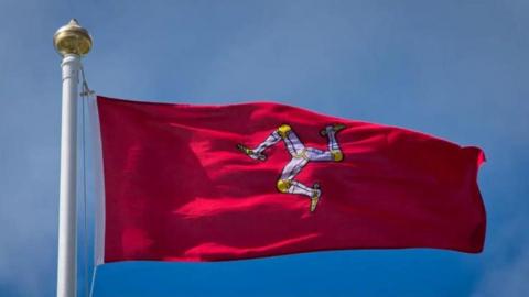 A Manx flag, which is red and features a white and yellow triskelion, blowing in the wind. It is attached to a white flag pole and the the sky in behind it is blue.