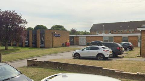 A Google Streetview image of a brown brick library building on a street with grass and trees in front of it. A few cars are parked and flats are in the background.