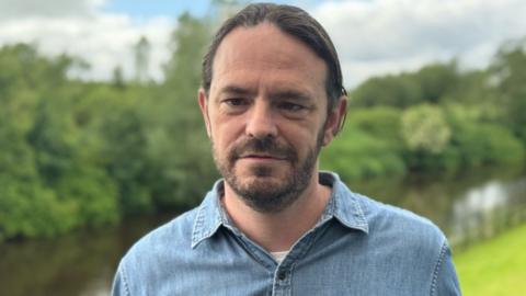 A man with dark hair and beard, he is wearing a denim blue shirt