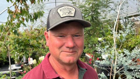 A man has a grey baseball cap on his head and a mauve open collar t-shirt top on, behind him are plants inside a polytunnel.