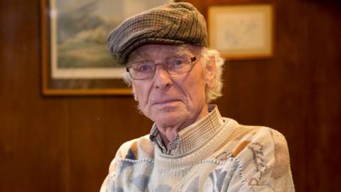 Jimmy Martin looking directly at the camera. He is wearing a multicoloured jumper and has his hands folded across his stomach. He is wearing a multicolured light bunnett and is standing in front of a wood background. A blurred fruit machine is on the right of the picture.