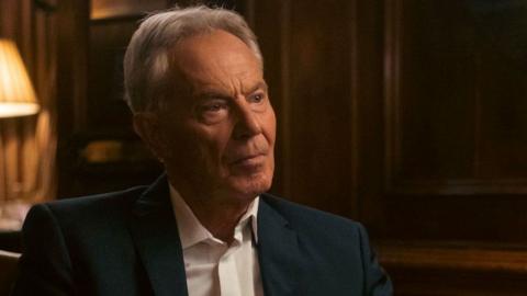 Sir Tony Blair sitting in a dimly-lit room, wearing no tie with his shirt's top button undone. He's looking to the side, off-camera. A lamp is lit behind him on the table