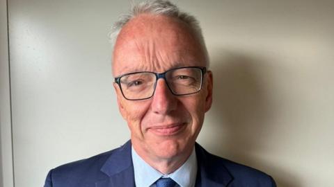 A man with greying hair, glasses and a navy blue suit smiles at the camera in an office.