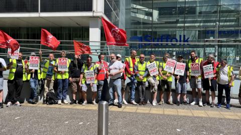 A line of people, most wearing hi-vis jackets. They are holding flags and signs. They are in front of a glass building.