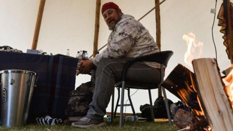 Pat King sits in a teepee during a sit-in protest at the Alberta Legislative Grounds. He wears jeans, a beige camouflage sweatshirt and red baseball cap.  