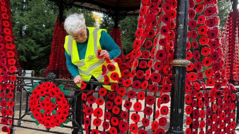 A woman wearing a bright yellow safety vest examines damage to netting displaying red poppies on a black metal bandstand in a park. The poppies hang in vertical rows and form a large circular arrangement on the railing.