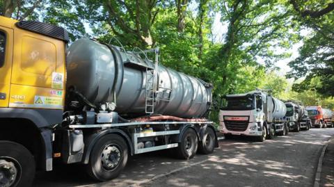 Tankers nose to tail along a tree-lined road.