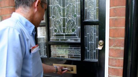 A Royal Mail postman posting letters through the letterbox of a black front door to a red brick house. He is wearing a blue shirt and has grey hair.