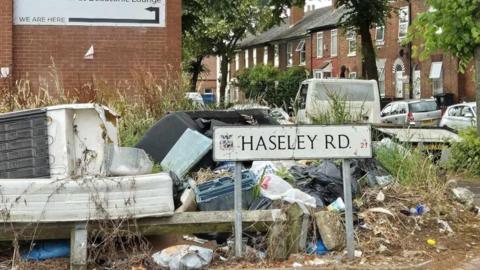 Dumped rubbish including a bed and packaging and wood with overgrown grass and weeds. In the middle is a road sign which says Haseley Road 