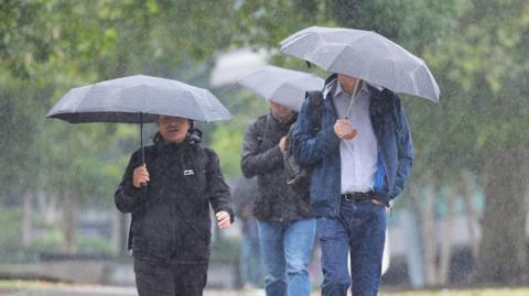 Three people walking outside in stormy weather conditions. They are all carrying umbrellas above their heads. It is raining.