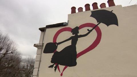 Mural shows black silhouette of Mary Poppins holding an umbrella surrounded by a red heart against a cream coloured wall