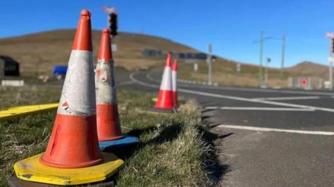 Orange and white traffic cones stand along the roadside. In the background, a road  can be seen winding around a hill, with tram lines on the far left side. It is a clear day, with blue sky overhead.