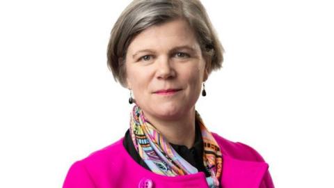 A woman with short grey hair and a bright pink coat, with a colourful silk scarf over the top.