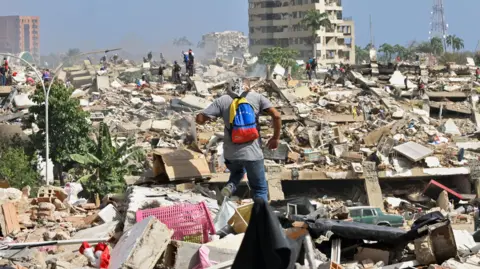 A man with a Venezuelan flag-themed backpack walks amid rubble of collapsed buildings after earthquakes hit the country, in La Guaira, Venezuela