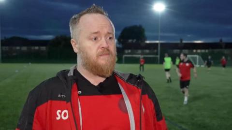 Sean O'Doherty has short hair and a beard. He is stood at the side of a floodlit football pitch. He is wearing a red and black t-shirt and a red and black tracksuit top.