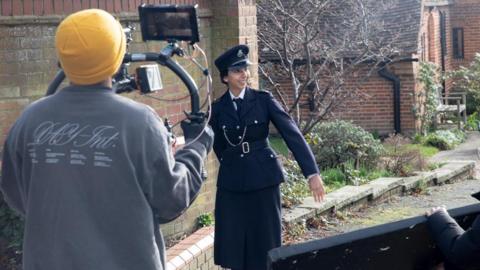 A woman acting as a police officer being filmed by a man in a yellow beanie with a big camera attached to a right. The actress is smiling and walking along the road.