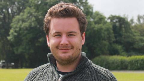 A close-up of Thom Campion smiling as he looks directly into the camera. He has brown hair, brushed away from his face, and a thin beard. He is wearing agreen woolly top and standing in a field with trees behind him.