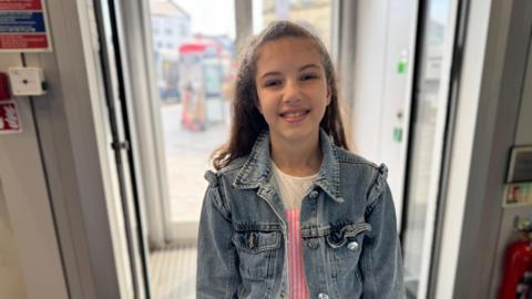 A girl in a denim jacket smiles at the camera by a door