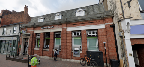 A high street bank brown-bricked building. Silver lettering reads 'Lloyds Bank'. A woman is at the cashpoint withdrawing money. A man in yellow hi-vis is walking past. 