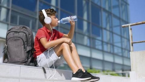 School boy drinks water with backpack outside school