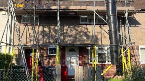 A terraced house with scaffolding on it. On the first floor, the scaffolding looks to be blocking the upstairs windows. The front door of the house is white. In the foreground, there is a garden gate.