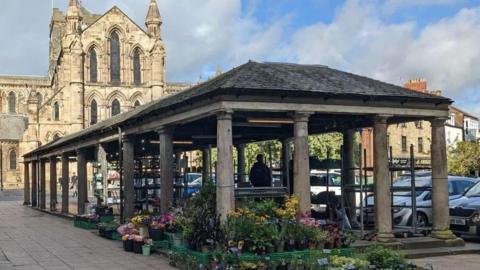 The Shambles is a Grade-II listed structure which dates back to 1766. It is a stone structure with a series of pillars holding up a tiled roof. It is an open structure without walls or windows and outside it are trays of potted plants. Part of Hexham Abbey, a huge church, is visible in the background. A row of cars are parked on the far side of The Shambles.