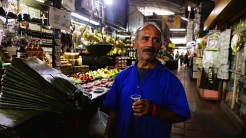 A man in a Caracas market speaks while holding a glass of coffee
