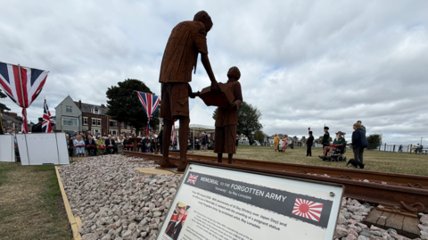 The sculpture, which is made from weathered corten steel and is now a dark brown colour, has been fixed to the ground and is surrounded by a bed of railway ballast. The sculpture shows a tall man, Len Gibson passing down a banjo that he played to prisoners to a young Burmese boy after the liberation of the camps. In the background, commemorations are taking place on a section of field with members of the public and veterans honouring those who died. 