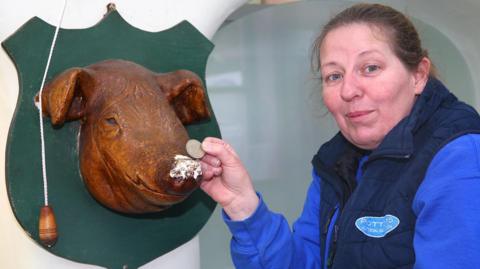 Kat Lister is standing beside a mounted wooden pig’s head sculpture attached to a shield-shaped green plaque on a wall. She holds a coin up to the pig’s snout, which has a small, worn or chipped patch on the surface. A pull cord with a wooden handle hangs next to the mounted sculpture on the left. Kat is wearing a blue long-sleeved top under a dark padded vest with a logo on the chest. The background shows part of a curved wall or doorway.