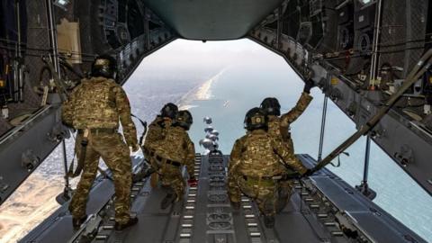 Airmen in khaki uniform watch from the open rear of an airplane as supplies are dropped by parachute 