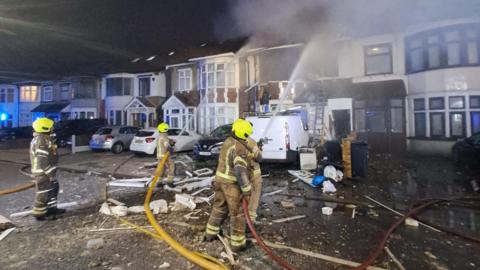 Firefighters in yellow helmets are working at the scene of a house fire at night, with smoke billowing from a damaged terraced house. Debris is scattered across the street, and fire hoses are in use.