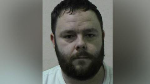 Mugshot of Binns. He has short dark hair and a thick dark beard and is wearing a white t-shirt.