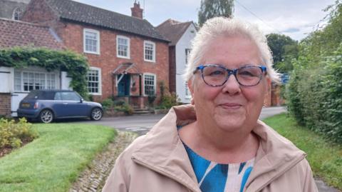 Pam Powell stands in a village lane with neat grass verges to either side and a red-brick cottage and white house in the background. She has short white hair, wears blue glasses and a cream-coloured coat and smiles towards the camera.